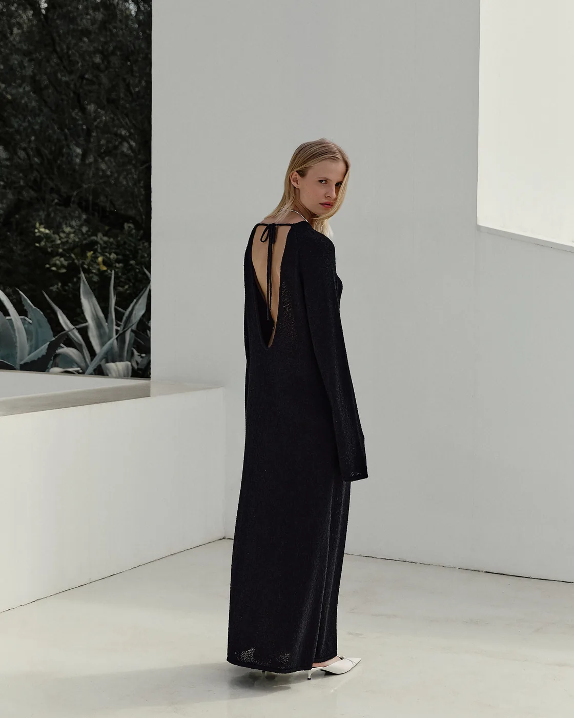 Model in a long black open-back dress in a bare white room.