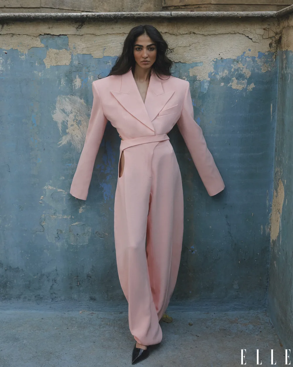 Model in a pale pink tailored suit against a weathered blue wall.